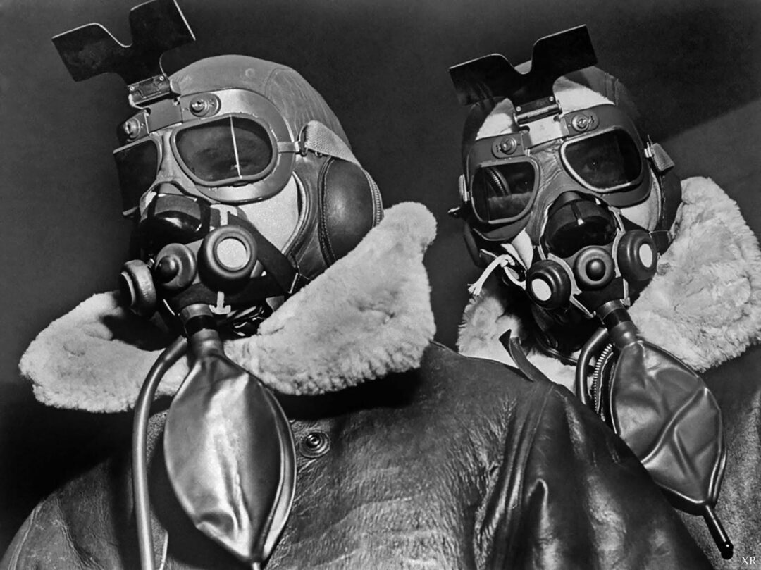 Two pilots wearing heavy leather flight jackets, goggles, oxygen masks, and headgear stand side by side, looking forward. The image is black and white, giving it a vintage, historical appearance.