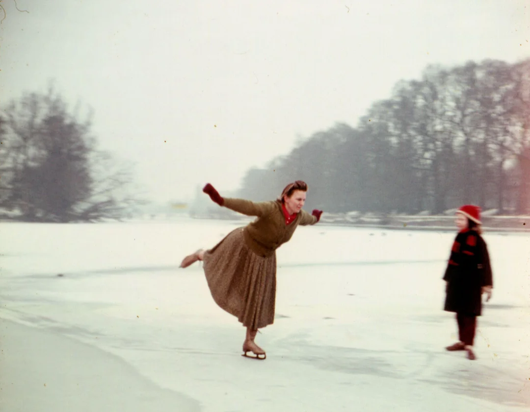A woman ice skating outdoors on a frozen pond, balancing on one foot with arms extended, while a child in a red hat and coat watches. Snow and bare trees are visible in the background.