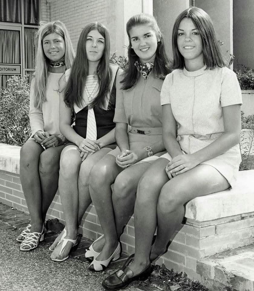 Four young women in 1960s-era clothing sit on a low brick wall outside a building, smiling at the camera. Three wear short dresses with scarves or ties, and one wears a vest and tie over a blouse.