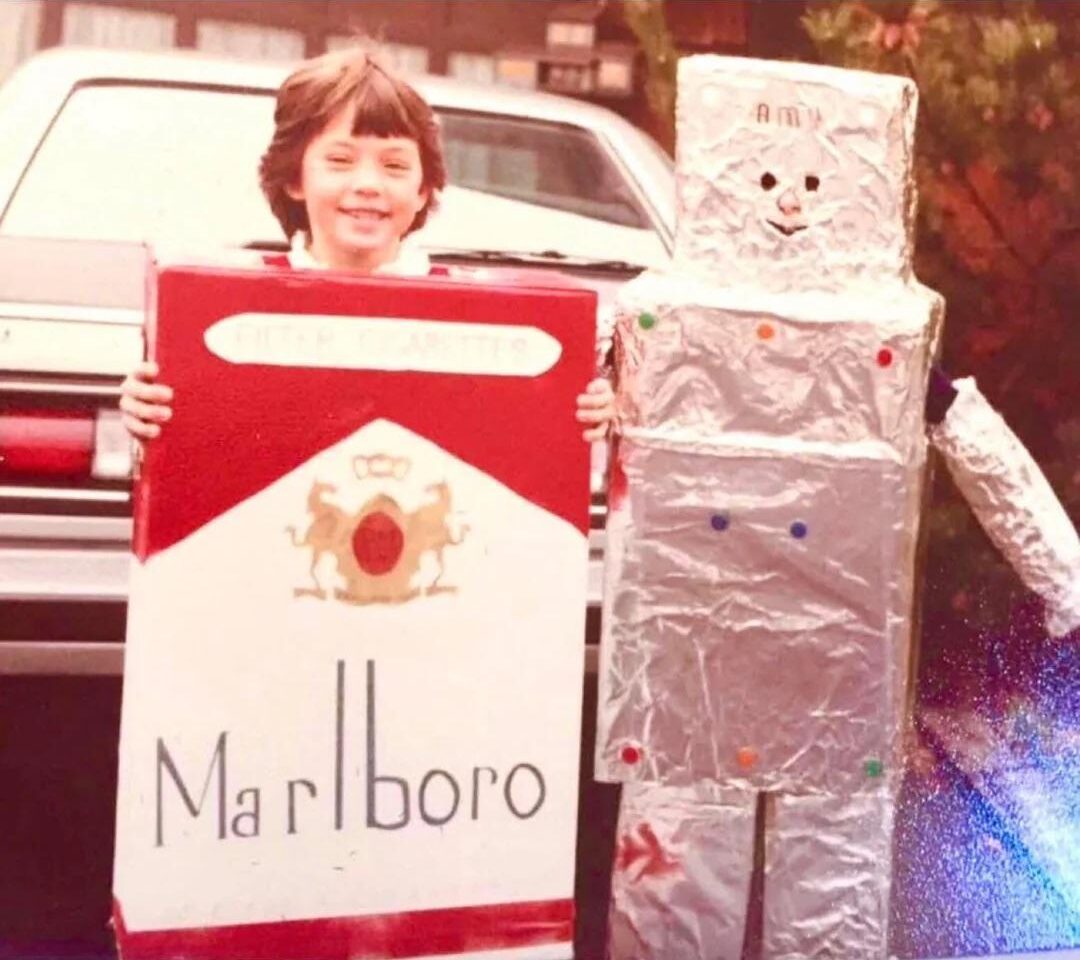 A child smiles while wearing a homemade Marlboro cigarette box costume, standing next to another child in a homemade robot costume made of foil-covered boxes. The text reads, “Homemade costumes in the 80s were legit.”