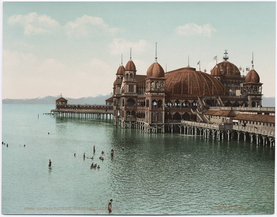 People swim in the water near a large, ornate pavilion on a pier extending into a lake, with mountains visible in the background under a clear sky.