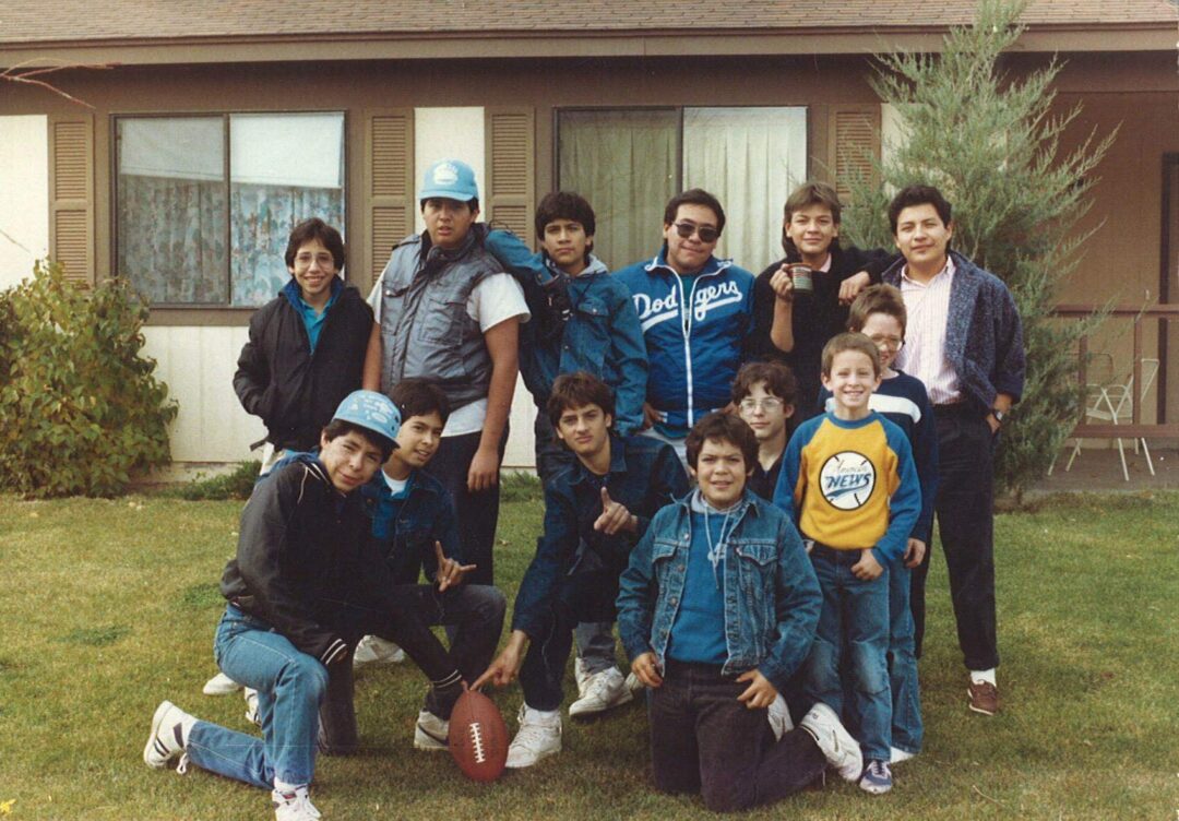 A group of boys and men pose outside a house on grass, some wearing jackets and hats. One person in front kneels by a football. Everyone is smiling, with a tree and patio chairs in the background.