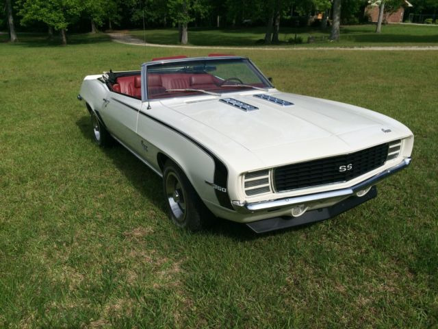 A white classic convertible car with a black grille and “SS” badge, parked on green grass. The car has red interior seats, chrome accents, and is surrounded by trees in the background.