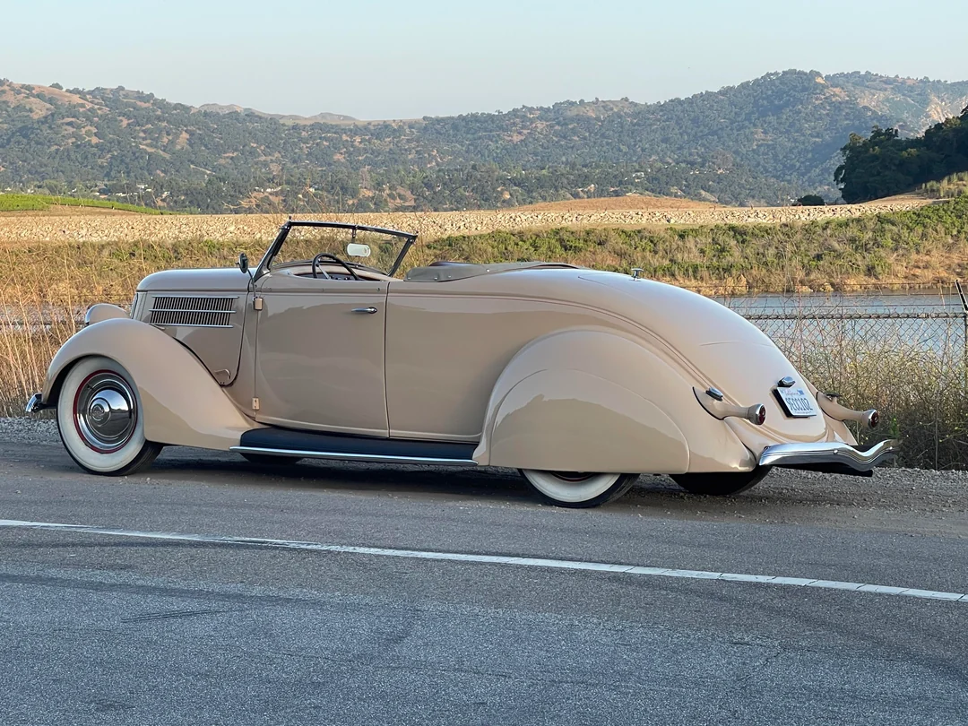 A vintage beige convertible car with whitewall tires is parked on the roadside. The background features a river, hills, and greenery under a clear sky.