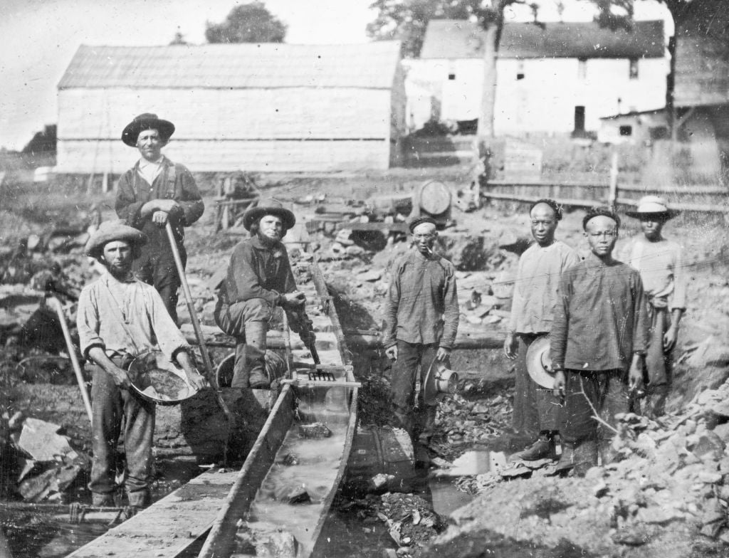A group of men, including Chinese laborers, stand by a sluice in a rocky area during the Gold Rush era, holding tools and gold pans, with wooden buildings and trees in the background.