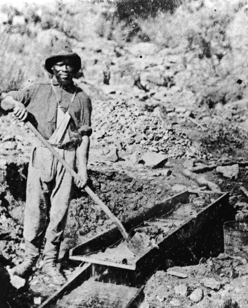 A man wearing a hat and rough work clothes stands outdoors, holding a shovel and working at a gold sluice, with rocks and dirt surrounding him in a rugged landscape.