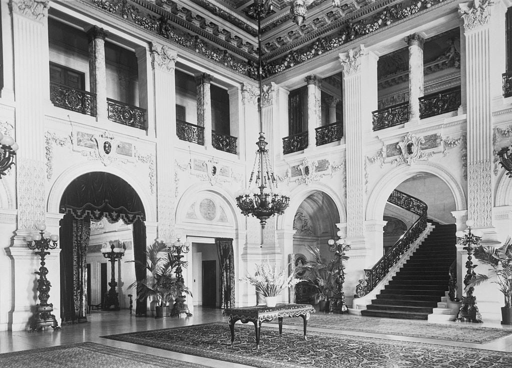 Ornate, grand foyer with tall columns, a curved staircase, intricate ceiling, decorative moldings, chandeliers, plants, and a large rug covering the marble floor. A central table with a flower arrangement stands in the middle.