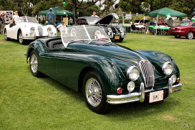 A dark green vintage convertible sports car is parked on grass at a classic car show, with several other classic cars, people, and tents visible in the background.