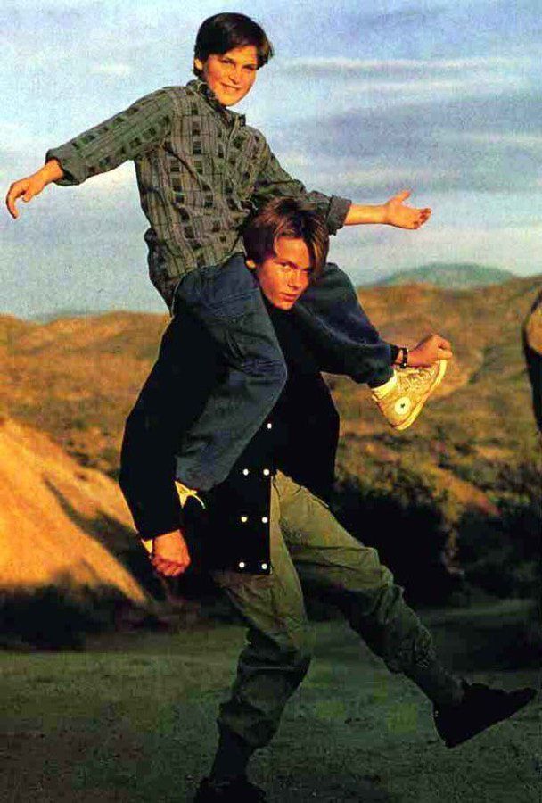 A young boy sits playfully on the shoulders of a teenage boy outdoors, both smiling. They are casually dressed, with hills and blue sky in the background.