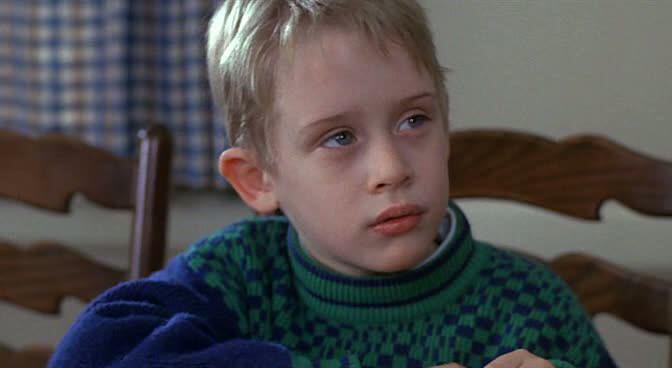 A young boy with blonde hair, wearing a green and blue checkered sweater, sits at a wooden table looking slightly to the side with a thoughtful expression. A checkered curtain is in the background.
