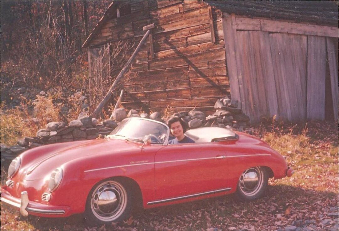 A person smiles while sitting in a red vintage convertible parked on grass in front of an old wooden barn, surrounded by autumn foliage and a stone wall.