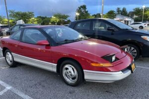 A red and silver two-door car with a pop-up headlight raised, parked in a lot near other vehicles on a cloudy day. Trees and lamp posts are visible in the background.