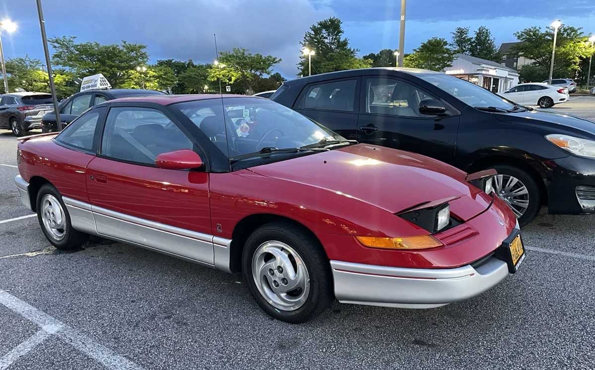 A red and silver two-door car with a pop-up headlight raised, parked in a lot near other vehicles on a cloudy day. Trees and lamp posts are visible in the background.