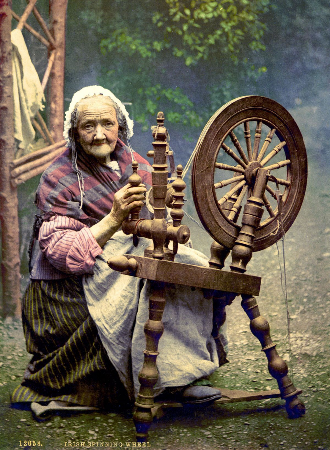 An elderly woman wearing traditional clothing sits outdoors, spinning yarn at an old-fashioned wooden spinning wheel. Lush greenery surrounds her, and she looks toward the camera.