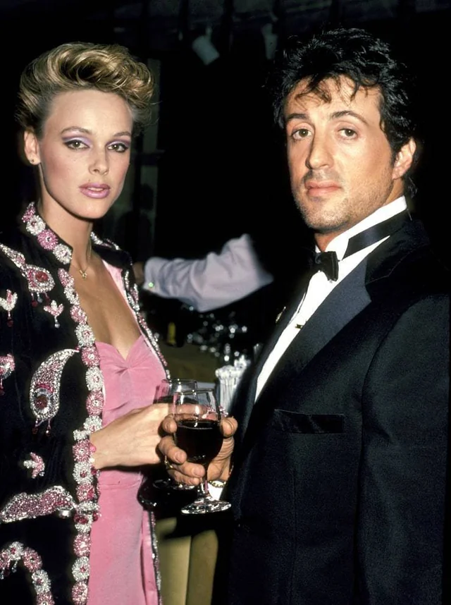 A woman in an ornate, embellished jacket holds a glass of wine beside a man in a black tuxedo. Both face the camera at a formal event with a dimly lit background.