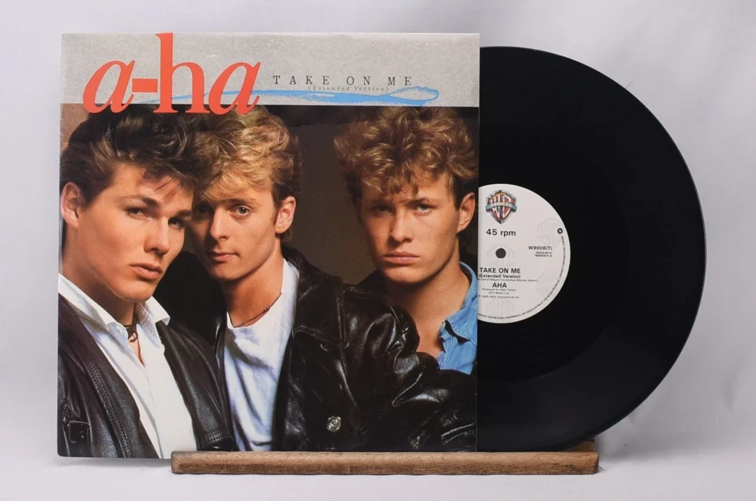 Vinyl record of a-ha's "Take On Me" with its cover showing three men in leather jackets. The album is propped up next to the black record, and "ATLAS RECORDS" is displayed below.