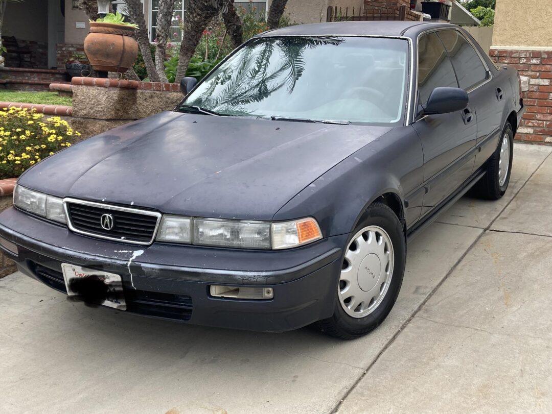A dark blue, older-model Acura sedan is parked in a residential driveway, with some visible scratches on the front bumper and a small dent near the right headlight.