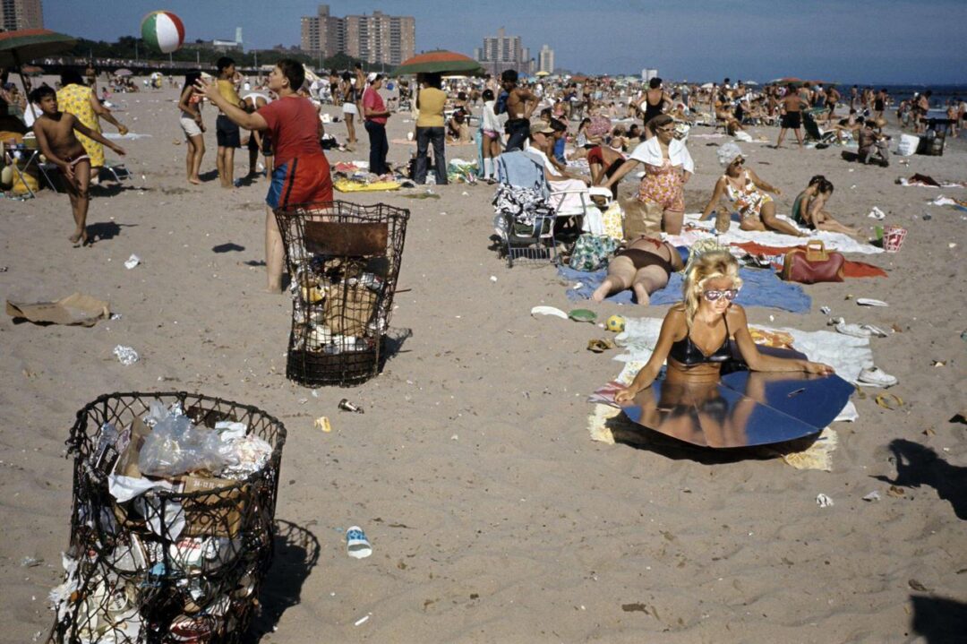 A crowded beach scene with people sunbathing, playing, and relaxing. The sand is scattered with litter and trash bins, while a woman in a bikini uses a reflective sunshield. Tall buildings are visible in the background.