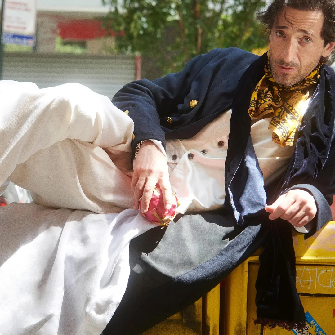 A man in vintage-style clothing, including a navy coat, cream trousers, and a yellow scarf, lounges on his side atop a yellow object outdoors, holding a pink flower and looking at the camera.