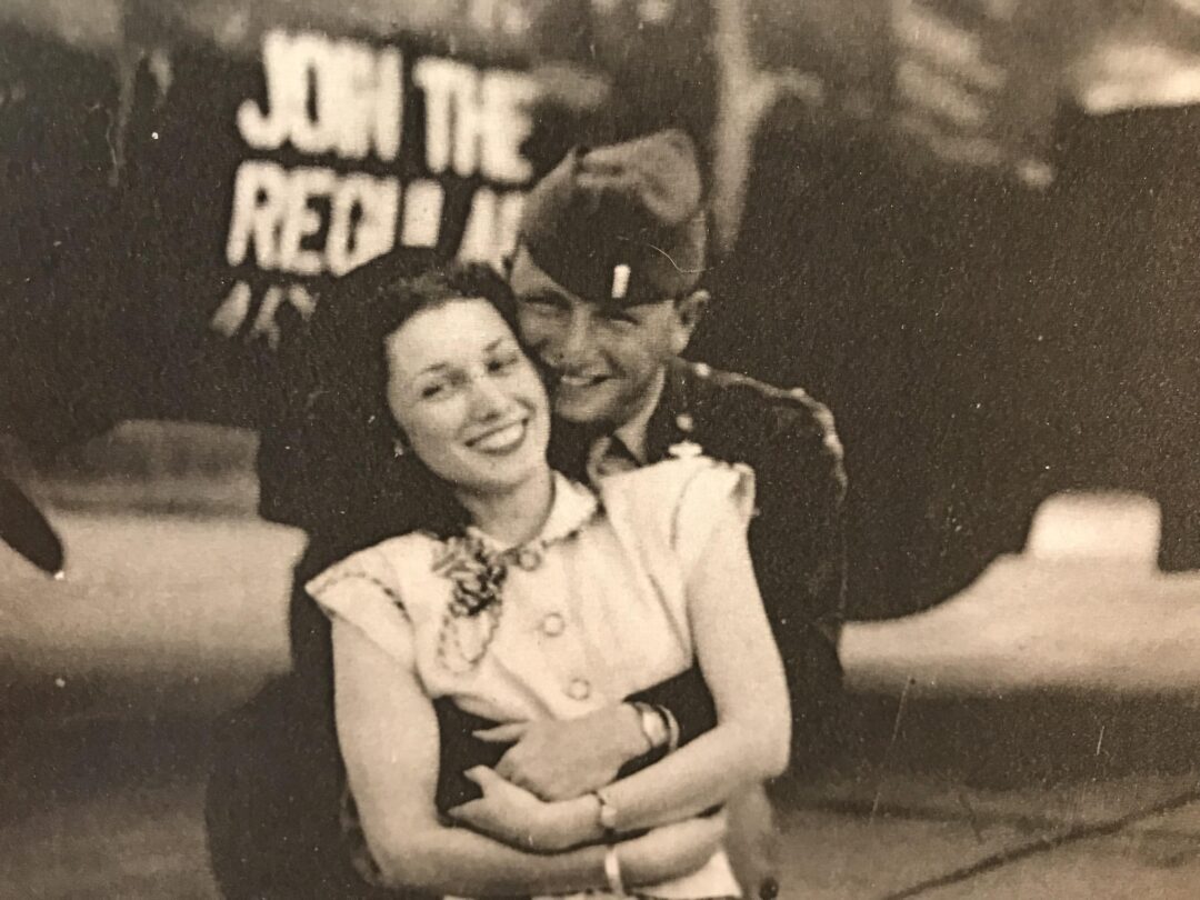 A smiling woman is embraced from behind by a man in a military uniform. Both appear happy, and a blurred sign in the background reads "JOIN THE RED ARMY." The photo is in black and white.
