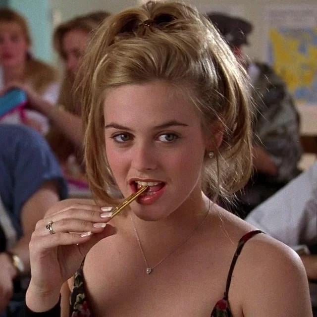 A young woman with blonde hair in a high ponytail bites the end of a pencil while sitting in a classroom. She wears a floral top and a necklace, looking thoughtfully ahead. Other students are visible in the blurred background.