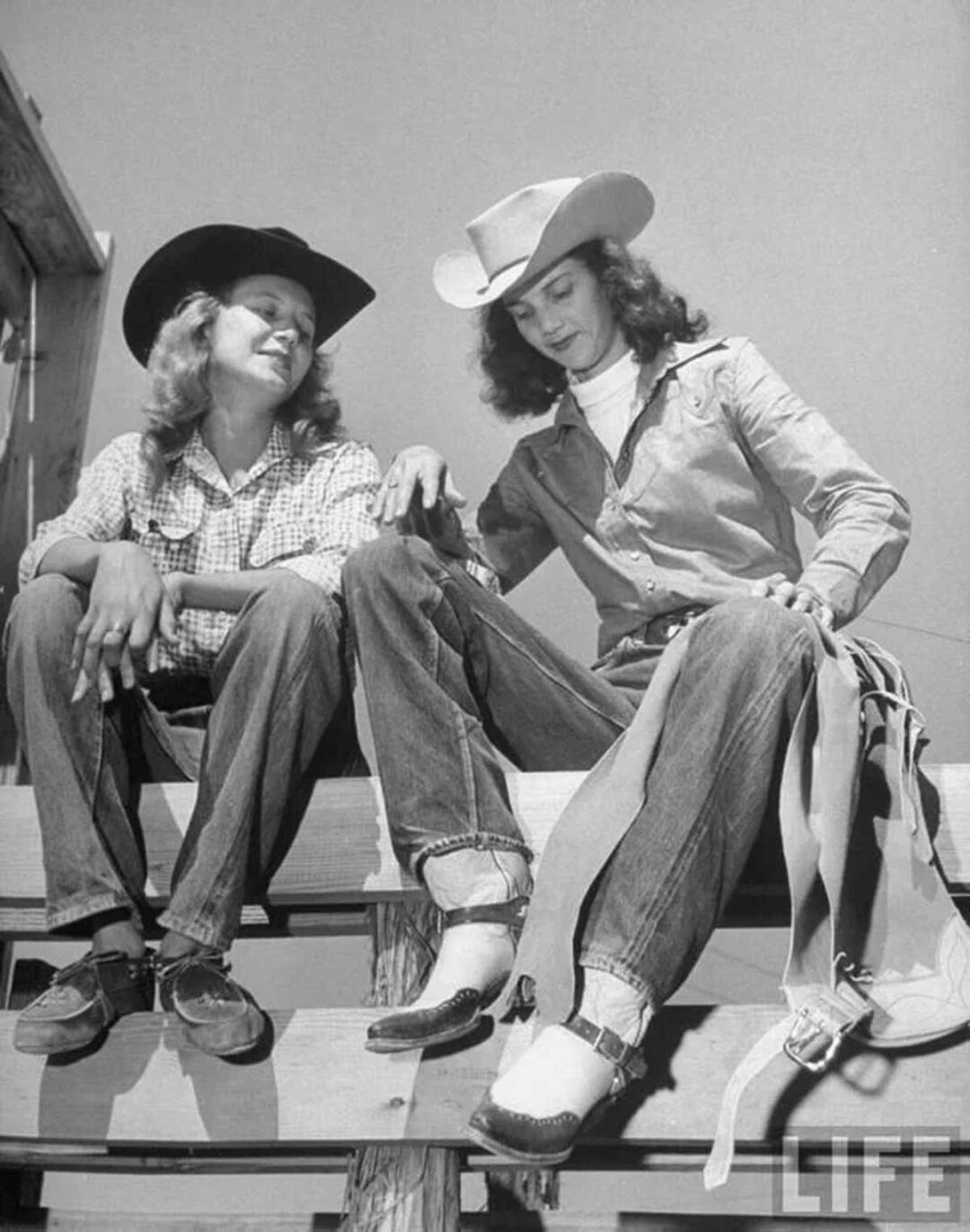 Two women wearing cowboy hats, plaid shirts, and jeans sit on a wooden fence, smiling and talking. One holds a saddle. The image is black and white and features a LIFE magazine watermark.