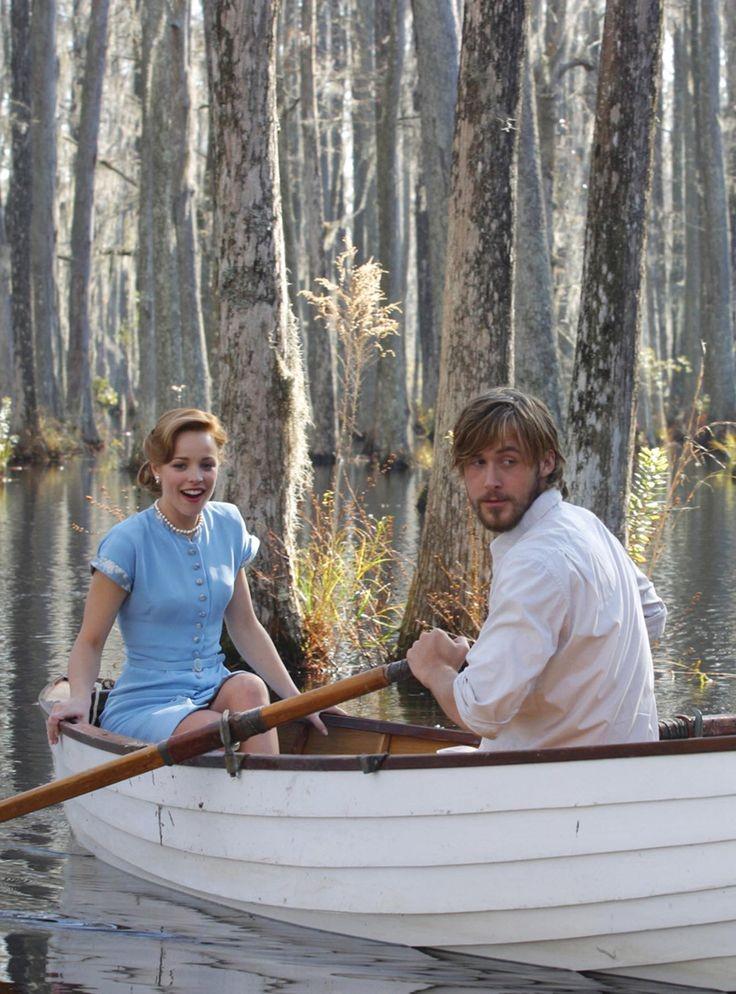 A man and woman sit in a white rowboat on a calm lake surrounded by tall trees. The woman wears a light blue dress and is smiling, while the man, in a white shirt, is rowing and looking toward the camera.