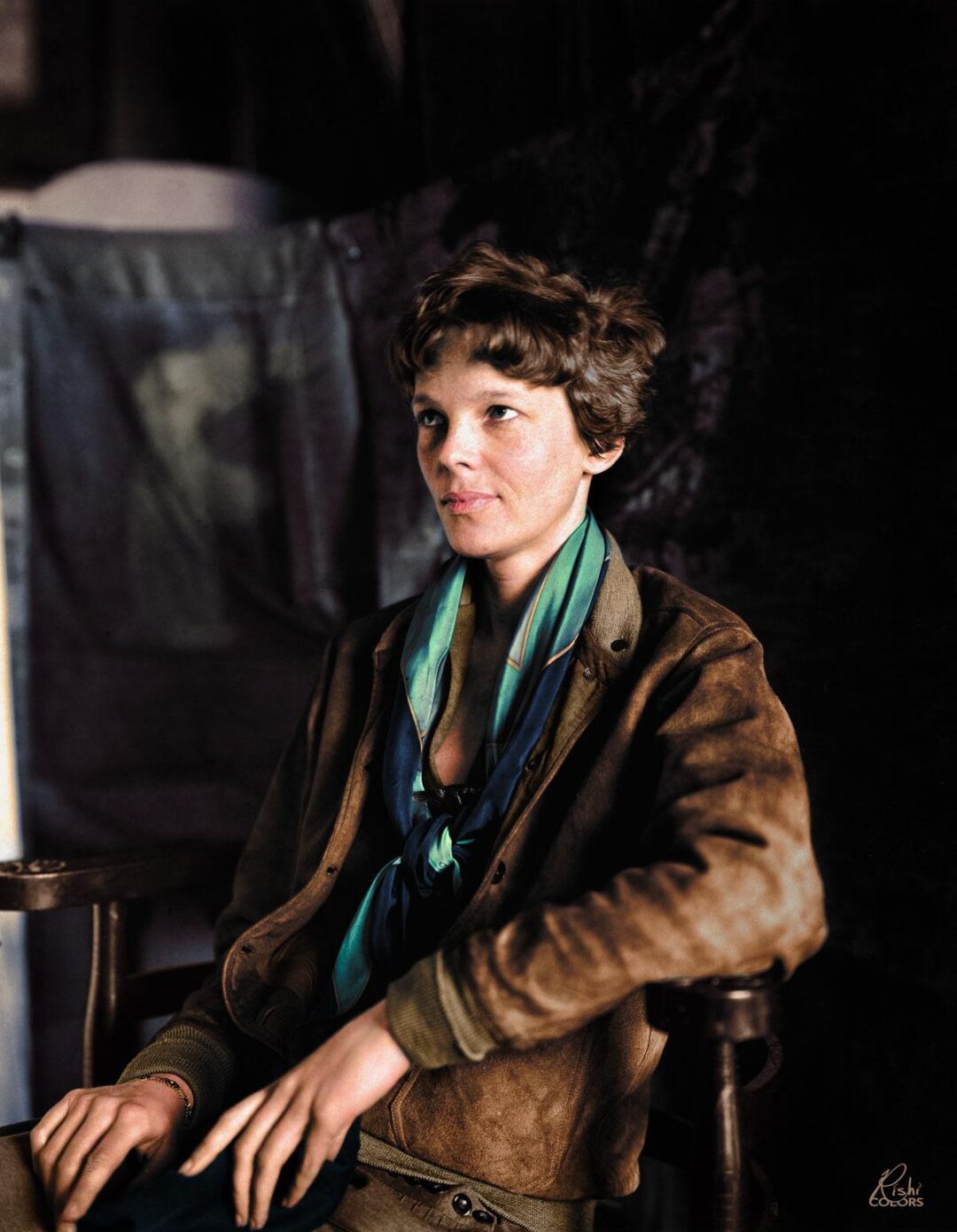 A woman with short brown hair sits on a wooden chair, wearing a brown leather jacket over a green shirt and a blue-green scarf, with a neutral expression and one hand resting on her lap. The background is dark and blurred.
