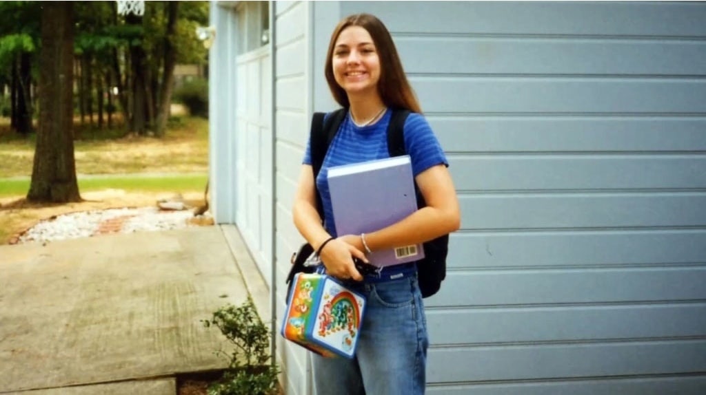 A smiling teenage girl stands outside by a driveway, wearing a blue striped shirt and jeans. She holds a binder and a colorful lunchbox, with a backpack on her shoulders. Trees and a garage are in the background.