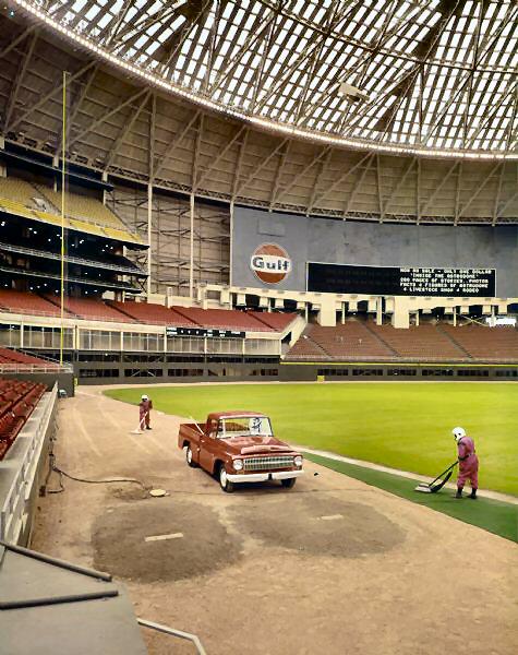 A color photo of an empty indoor baseball stadium with a large scoreboard, a Gulf Oil advertisement, a vintage red pickup truck parked near the outfield, and two workers tending the grass.