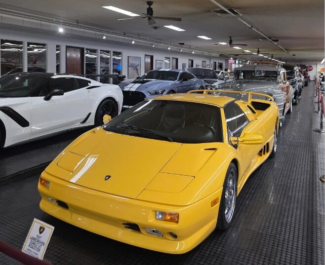 A yellow Lamborghini Diablo is displayed in a car museum, surrounded by other luxury vehicles, including a white sports car and a classic car, all lined up in a well-lit indoor showroom.