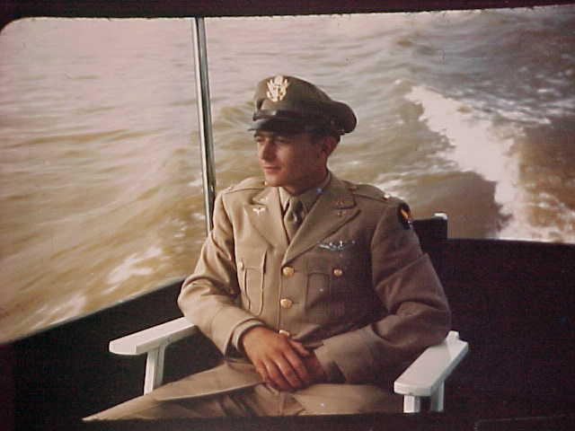 A young man in a tan military uniform and cap sits on a white chair aboard a boat, with water and waves visible in the background.
