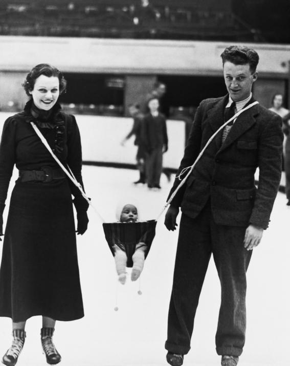 A woman and man stand on an ice rink, smiling, holding straps of a sling that carries a bundled baby between them. People skate in the background. The image is black and white.