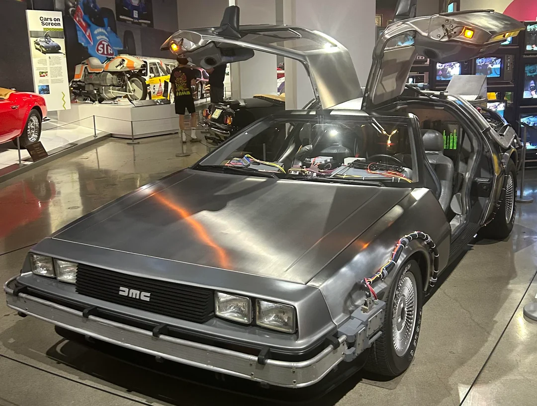A silver DeLorean car with gull-wing doors open, modified with sci-fi elements from "Back to the Future," is on display in a museum with other classic cars and exhibits visible in the background.