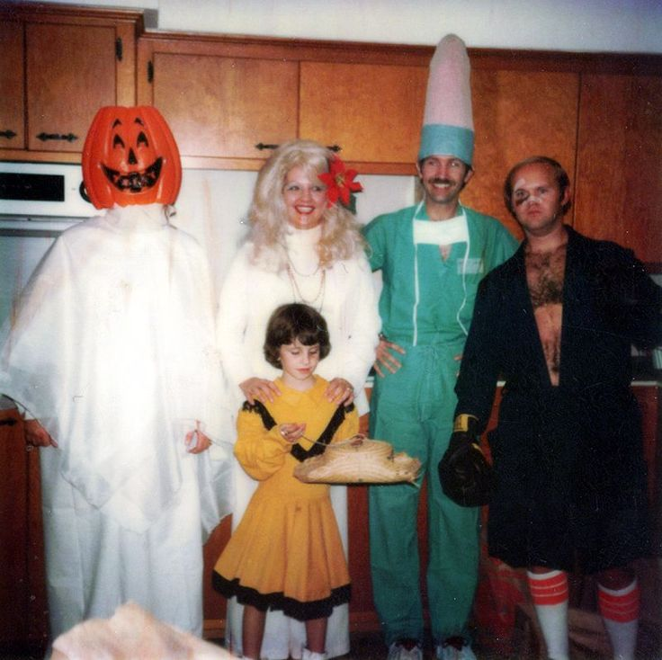 Five adults in unique costumes and one child pose in a kitchen. Costumes include a pumpkin head, a nurse with a tall hat, a boxer, a woman in white, and a child dressed in yellow holding bread.