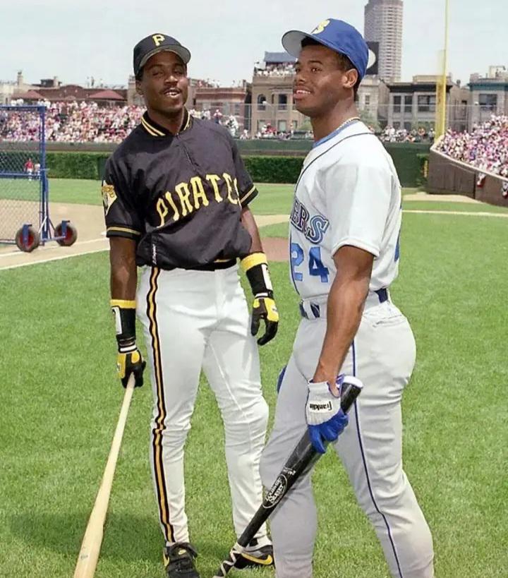 Two baseball players in uniforms stand on a field, each holding a bat. One wears a black Pittsburgh Pirates uniform, and the other wears a gray and blue Mariners uniform. A stadium and crowd are visible in the background.