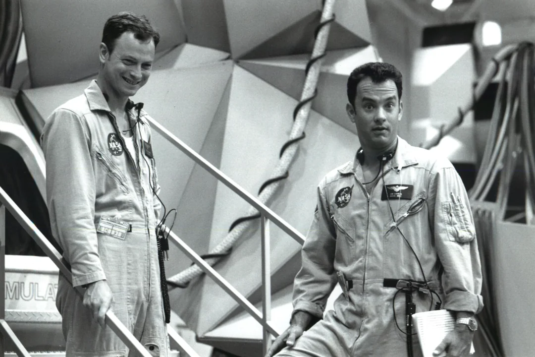 Two men in NASA astronaut jumpsuits stand in front of a geometric simulator set. One smiles while the other looks serious, holding a folder. The background features cables and futuristic structure details. Black and white photo.