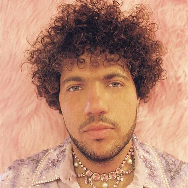 A person with curly hair and light-colored eyes poses against a pink, furry background. They wear layered necklaces and a lavender, beaded garment, looking directly at the camera with a neutral expression.