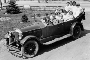 A vintage black-and-white photo shows a man driving an old-fashioned convertible car filled with at least twelve smiling children, seated close together. The car is on a dirt road with grass and trees nearby.