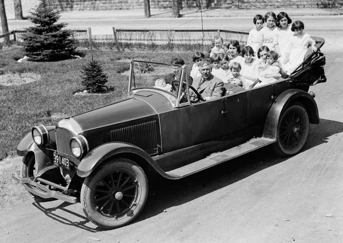 A vintage black-and-white photo shows a man driving an old-fashioned convertible car filled with at least twelve smiling children, seated close together. The car is on a dirt road with grass and trees nearby.