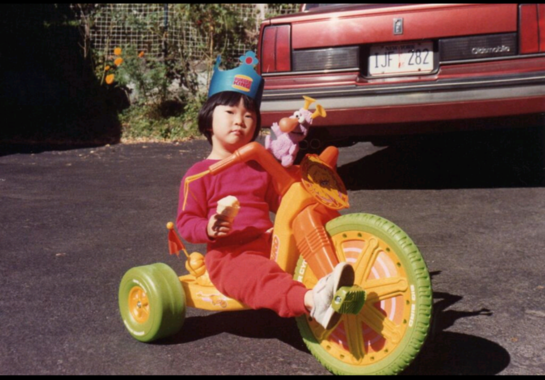 A young child wearing a crown and red clothes relaxes on a colorful tricycle, holding an ice cream cone. A stuffed animal is attached to the bike. A red car and greenery are in the background.