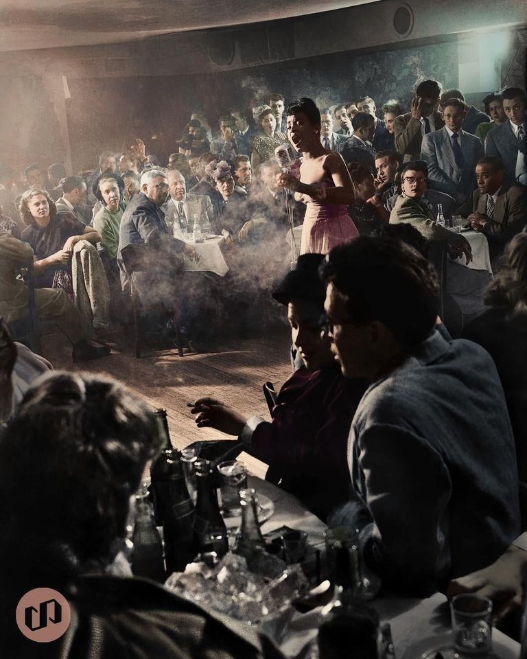 A female singer performs on stage in a crowded, smoky jazz club, surrounded by seated, well-dressed patrons at tables filled with drinks, all focused on her performance.