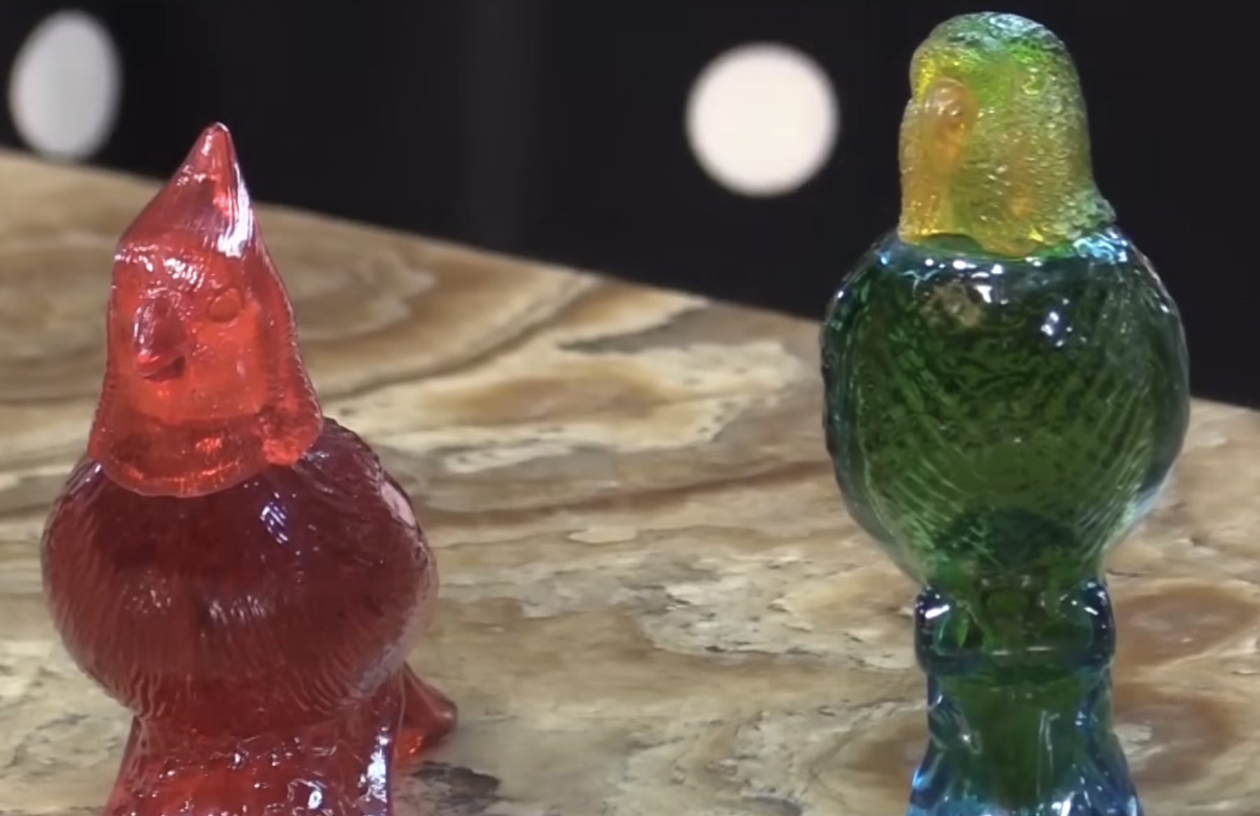 Two bird-shaped candies sit on a marble surface. One is a red cardinal, and the other is a green and yellow parrot. The background is dark with white spots.