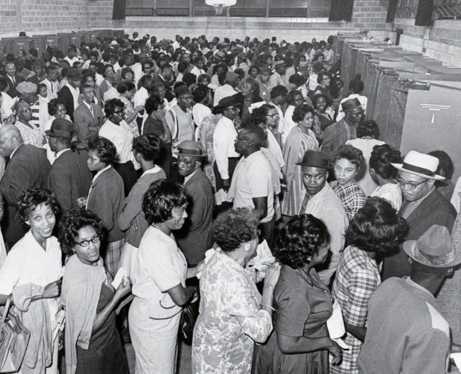 A large group of Black men and women stand in long lines inside a gymnasium, waiting to vote at curtained voting booths during the 1960s. People are dressed in formal and casual attire, some looking toward the camera.