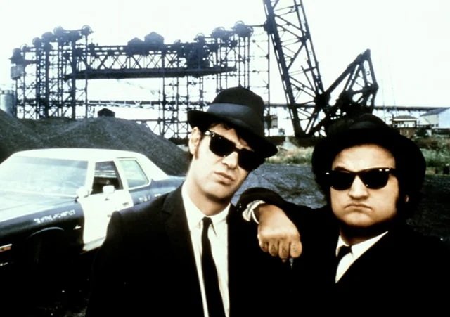 Two men in black suits, hats, and sunglasses stand in front of a car with industrial structures and cranes in the background. One man leans his arm on the other's shoulder.
