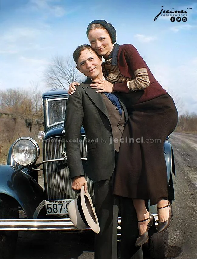 A man in a suit holds a hat while a woman in a burgundy and brown outfit sits on the car’s front bumper, hugging him. They pose in front of a vintage blue car on a rural road with bare trees and blue sky.
