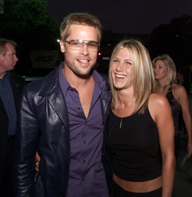 A man in a dark leather jacket and sunglasses stands smiling with a woman in a black sleeveless top, who is also smiling. They pose together outdoors at night, with other people and trees in the background.