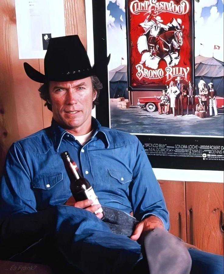 A man in a blue shirt and black cowboy hat sits indoors holding a beer bottle. Behind him is a movie poster for "Bronco Billy" featuring a cowboy on a horse.