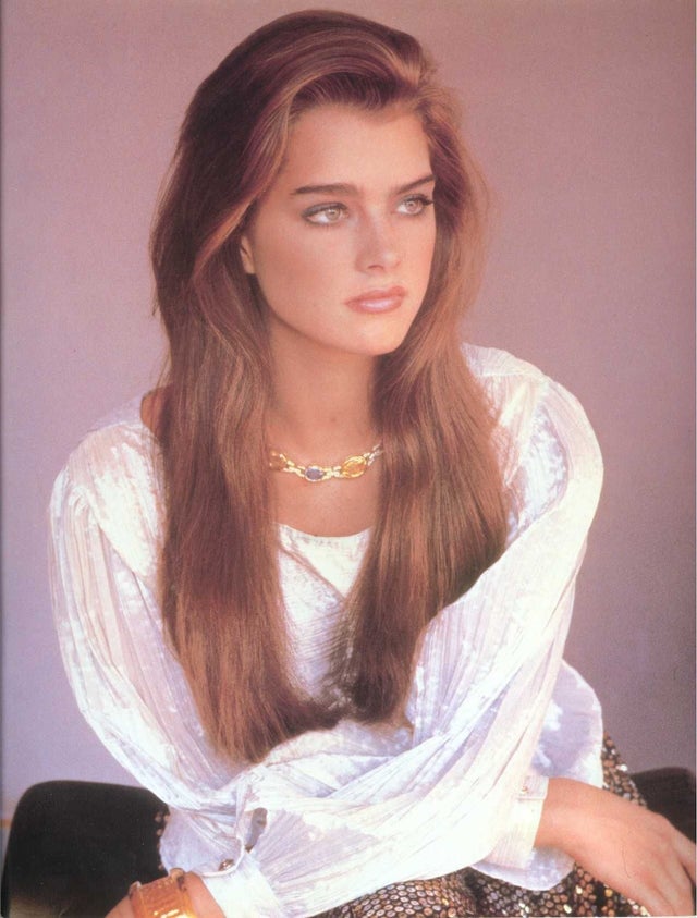 A woman with long brown hair sits looking to the side. She wears a white, textured blouse, a gold necklace, and a gold bracelet. The background is a soft lilac color.