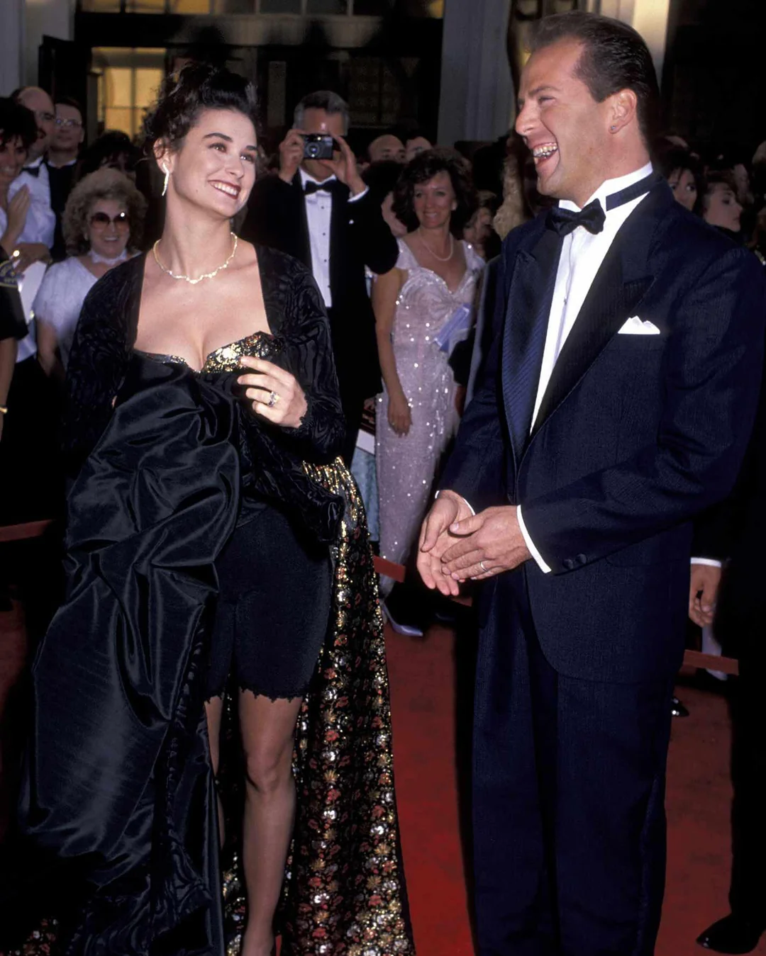 A woman in a glamorous black and gold gown smiles and looks at a man in a tuxedo as they stand on a red carpet, surrounded by people dressed formally and photographers in the background.